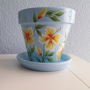 May include: A light blue terracotta flower pot with a painted design of yellow hibiscus flowers and green leaves. The pot has a matching saucer.