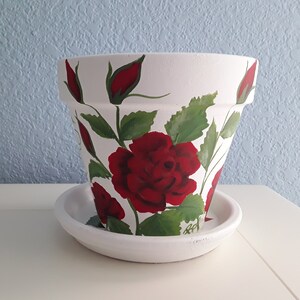 May include: A white ceramic flower pot with a matching saucer, featuring hand-painted deep red roses and green leaves wrapping around the exterior. The design includes several blooming red rose flowers and closed rose buds on stems.