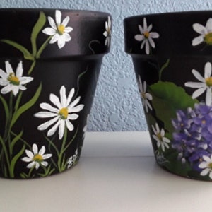 May include: Two black terracotta flower pots with white daisy and purple flower designs painted on them.