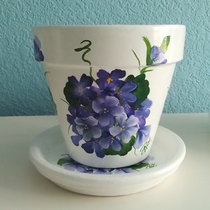 May include: White ceramic flower pot and matching saucer, hand-painted with a cluster of purple violets and green leaves. The pot has a wide rim and a curved shape. The saucer has a matching violet design.