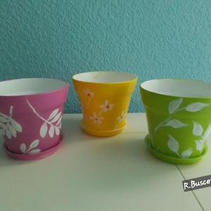 May include: Three painted flower pots, one pink with white leaf designs, one yellow with white flower designs, and one green with white leaf designs.