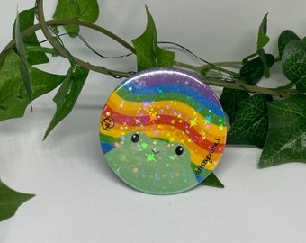 May include: A round, green button with a cartoon frog face and a rainbow design. The frog has a rainbow-colored hair and star accents. The button has the text "artsycristal".