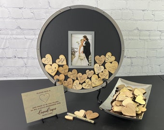 Peut inclure: Un livre d'or de mariage avec un cadre noir et une photo de la mariée et du marié. Le cadre est entouré de cœurs en bois avec une finition argentée. Les cœurs sont destinés aux invités pour qu'ils y signent leurs noms. Le texte sur le panneau indique "Veuillez signer un cœur et le déposer dans le cadre afin que nous nous souvenions toujours de nos proches qui sont venus."