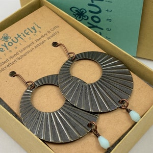 May include: Handmade, dark grey metal earrings with a sunburst pattern, displayed in a gift box. The earrings feature copper-coloured hooks and small, light blue teardrop-shaped beads.