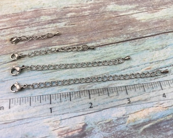May include: Four silver extender chains of varying lengths, measured in inches on a ruler. The chains are made of small, linked metal circles.