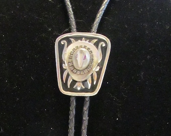 Puede incluir: Un corbatín bolo negro con un colgante plateado y negro con una piedra blanca. El colgante tiene un diseño decorativo con un motivo floral.