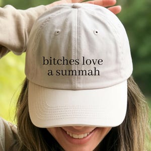 May include: A light beige baseball cap featuring the black text "bitches love a summah" embroidered on the front panel. The cap has a structured crown, a curved brim, and eyelets for ventilation, providing a classic and simple headwear design.