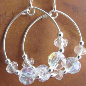 May include: Silver hoop earrings with clear, iridescent crystal beads. The earrings feature a simple, elegant design with the crystals adding sparkle and dimension. The crystals are various sizes and shapes, creating a unique look.