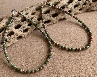 May include: A beaded necklace featuring green and pink unakite stones. The necklace has small, round beads with silver-colored spacers and a silver clasp. The beads are strung on a delicate chain, creating a natural and earthy aesthetic.