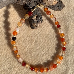 May include: A beaded bracelet featuring a mix of orange, red, and cream-colored gemstones. The bracelet is strung with small gold-colored beads and has a gold-colored clasp. The gemstones vary in size and shade, creating a warm, earthy aesthetic.