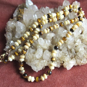 May include: A layered necklace featuring cream-colored and brown beads, interspersed with gold-colored accents. The necklace is displayed on a bed of white crystals against a maroon background. The beads have a polished finish.