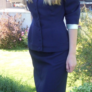 May include: A young woman wearing a navy blue suit with a white collar and a skirt. She is standing on a grassy lawn.