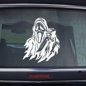 May include: A white vinyl decal of the Ghostface mask from the Scream horror film series, holding a knife. The decal is affixed to the rear window of a black Cadillac hearse.