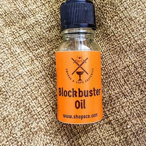 May include: A small, clear glass bottle with a black dropper top. The bottle is labeled "Blockbuster Oil" with a logo of crossed swords and cups. The label is orange with black text and the website address "www.shopsce.com".