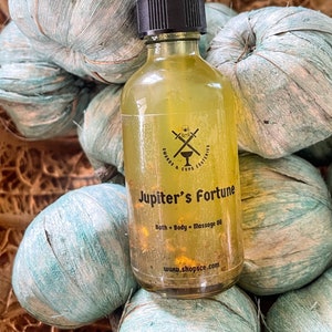 May include: A clear glass bottle with a black dropper top filled with a yellow liquid. The bottle has a black label with the text "Jupiter's Fortune" and "Bath + Body + Massage Oil". The label also includes a logo with crossed swords and a cup, and the text "Swords & Cups Esoterics". The bottle is sitting on a blue and brown textured surface.