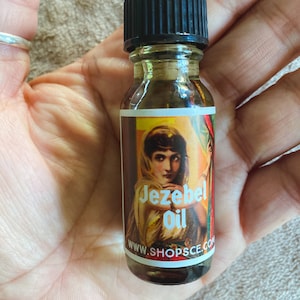 May include: A small glass bottle with a black cap, filled with a dark liquid. The label features a woman in a red and green dress with the text "Jezebel Oil" and a website address.