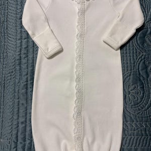May include: A white long-sleeved infant gown featuring a delicate white lace trim along the round neckline and down the center front opening. The garment has long sleeves with ribbed cuffs and an elasticized bottom hem for a secure fit.