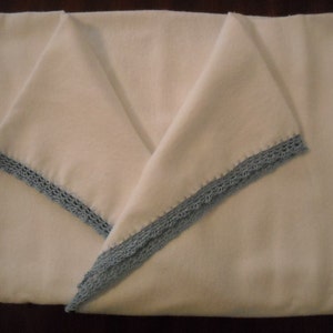 Puede incluir: Una manta blanca doblada con un ribete de ganchillo azul claro. La manta est&aacute; doblada en forma triangular, con el ribete visible a lo largo de los bordes. La manta parece estar hecha de un material suave.