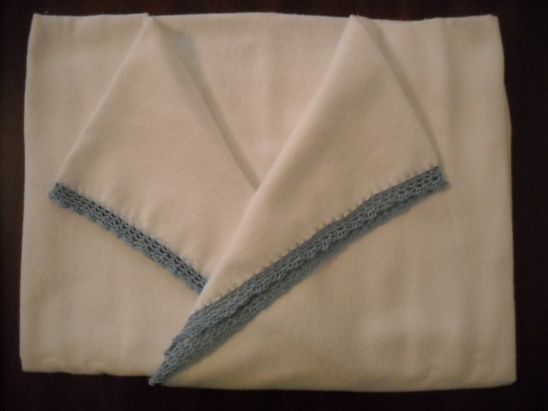 Puede incluir: Una manta blanca doblada con un ribete de ganchillo azul claro. La manta est&aacute; doblada en forma triangular, con el ribete visible a lo largo de los bordes. La manta parece estar hecha de un material suave.