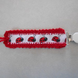 May include: Red crocheted baby pacifier clip with a white ribbon featuring a repeating pattern of ladybugs. The clip is attached to a white plastic ring.
