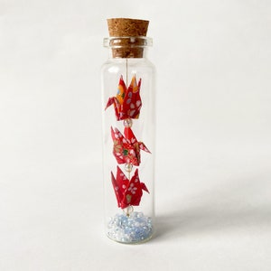 以下が含まれることがあります： コルク栓の付いたガラス瓶に、赤と黄色の折り鶴と白いビーズの糸が入っています。