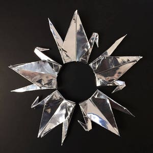 以下が含まれることがあります： 黒い背景に円形に配置された8羽の銀色の折り紙鶴