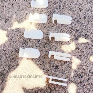 May include: A collection of white plastic curtain clips and hooks, arranged on a textured gray surface. The clips have various designs, including rounded and rectangular shapes. The image includes the text "&copy; HEARTSKIPGIFTS".