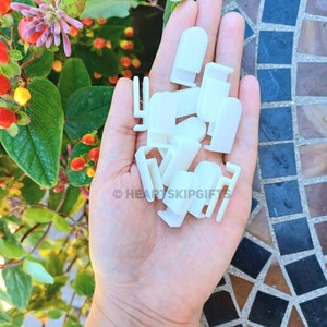 May include: A hand holding a collection of small, white plastic clips of various shapes. The clips are in focus against a backdrop of green foliage with red and orange berries and a mosaic tile surface. The text "HEARTSKIPGIFTS" is visible.