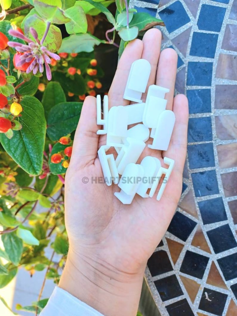 May include: A hand holding a collection of small, white plastic clips of various shapes. The clips are in focus against a backdrop of green foliage with red and orange berries and a mosaic tile surface. The text "HEARTSKIPGIFTS" is visible.