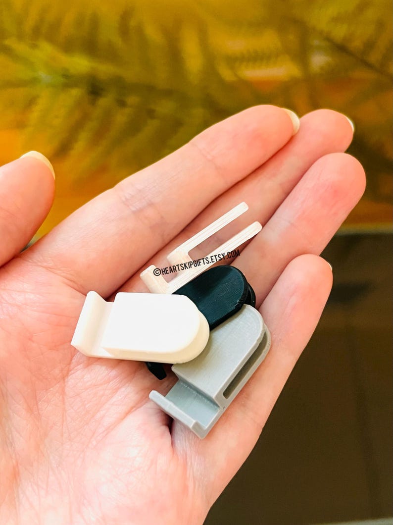 May include: A collection of small, plastic clips in various colors: white, black, and gray. One clip has two prongs. The text "@HEARTSKIPGIFTS.ETSY.COM" is visible on one of the clips. These clips are likely used for organizing or securing items.