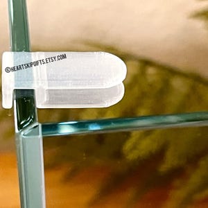 May include: A clear, plastic shelf support clip securing two glass panels at a 90-degree angle. The clip is white and has a rounded shape. The Etsy shop name "HEARTSKIPGIFTS.ETSY.COM" is printed on the clip.