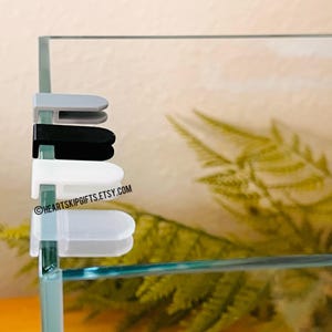 May include: A close-up of four aquarium lid clips in gray, black, white, and clear. The clips are attached to the glass of an aquarium. The text "HEARTSKIPGIFTS.ETSY.COM" is visible on the glass.