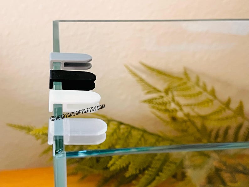 May include: A close-up of four aquarium lid clips in gray, black, white, and clear. The clips are attached to the glass of an aquarium. The text "HEARTSKIPGIFTS.ETSY.COM" is visible on the glass.