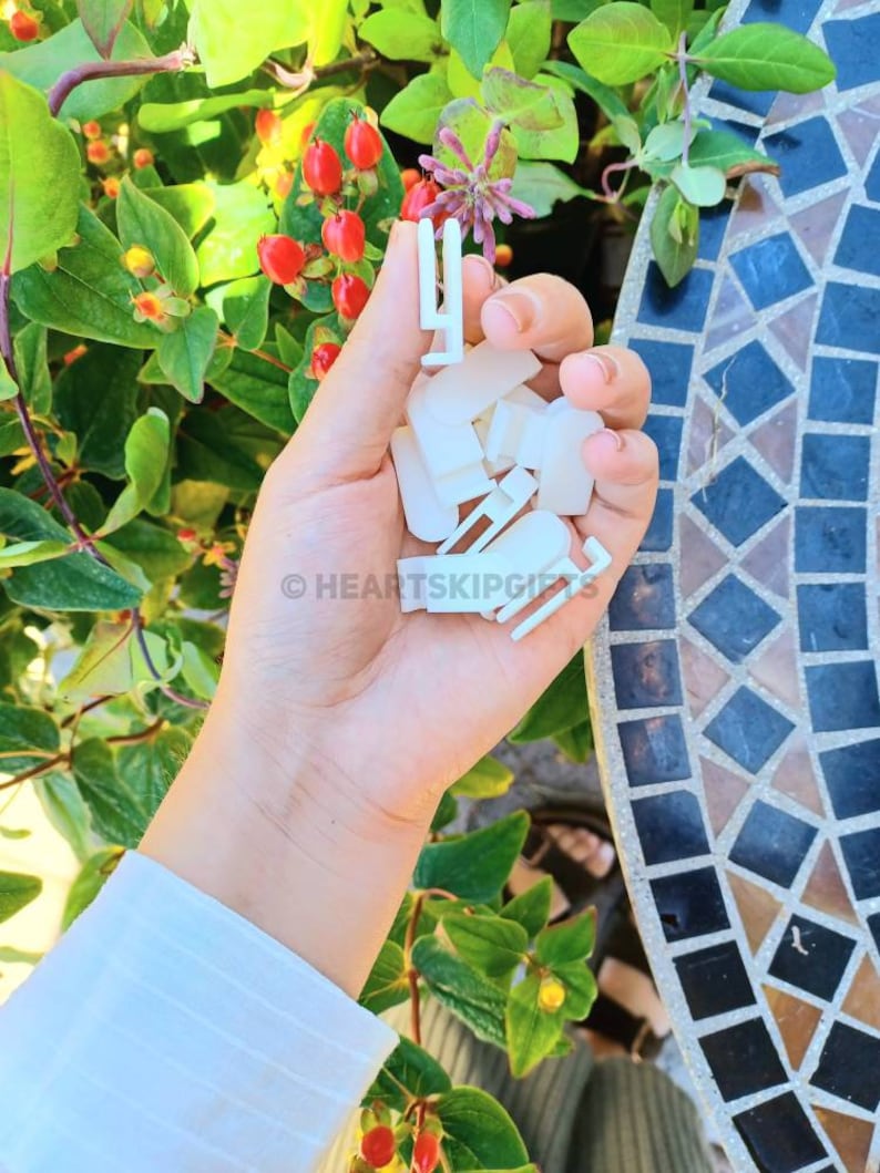 May include: A hand holding a collection of small, white plastic clips. The clips are various shapes and sizes, with some appearing to be designed for attaching items. The background features green foliage, red berries, and a mosaic tile surface.