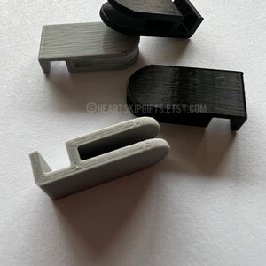 May include: Four 3D-printed plastic clips in black and gray. The clips have a rectangular shape with a curved top and a small hook on one side. The clips are arranged on a white surface. The Etsy shop name "HEARTSKIPGIFTS.ETSY.COM" is visible.