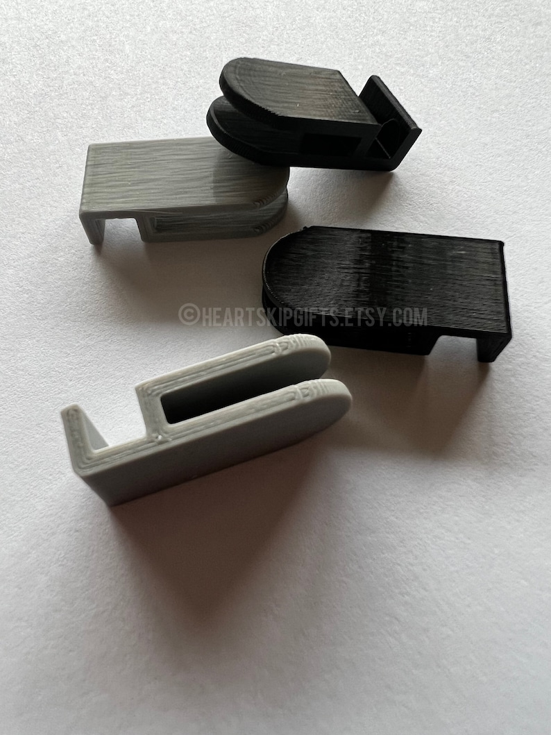 May include: Four 3D-printed plastic clips in black and gray. The clips have a rectangular shape with a curved top and a small hook on one side. The clips are arranged on a white surface. The Etsy shop name "HEARTSKIPGIFTS.ETSY.COM" is visible.