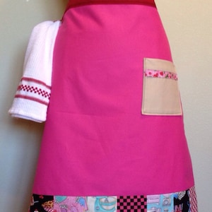 May include: A bright pink apron with a red waistband and a beige pocket with a cherry-patterned trim. The bottom of the apron features a patchwork design with various kitchen-themed patterns and checkered squares. A white towel with red accents hangs on the side.