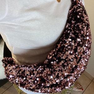 Peut inclure: Un sac banane en sequins rose doré avec une sangle noire et des détails de chaîne. Le sac est en forme de croissant et porté en bandoulière. Un haut blanc et un collier en or sont également visibles.