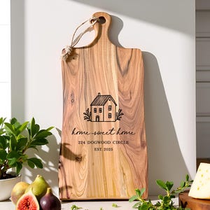 May include: A wooden cutting board with a handle and rope loop. The board features the text "home sweet home", a house graphic, and address details. The board is surrounded by figs, cheese, and greenery.