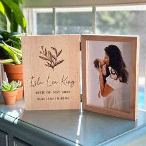 May include: A wooden photo frame with a light wood finish. The frame has a hinged front that opens to reveal a photo of a mother and baby. The front of the frame is engraved with the text "Isla Lea King", "26th of May 2023", "6 lbs 15oz | 6:43PM" and a floral design.