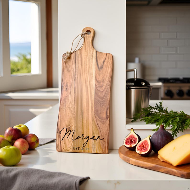 A personalized cutting board with the name "Morgan" and "Est 2025" displayed elegantly near a window with fruit and cheese.