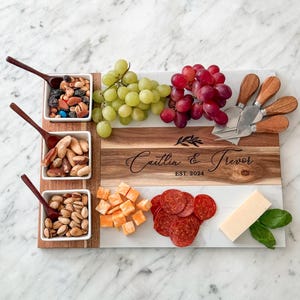 May include: A white marble and wood serving board with four small white bowls filled with snacks. The board has a personalised inscription that reads "Caitlin & Trevor EST. 2024". The board is surrounded by grapes, cheese cubes, pepperoni slices, and basil leaves.