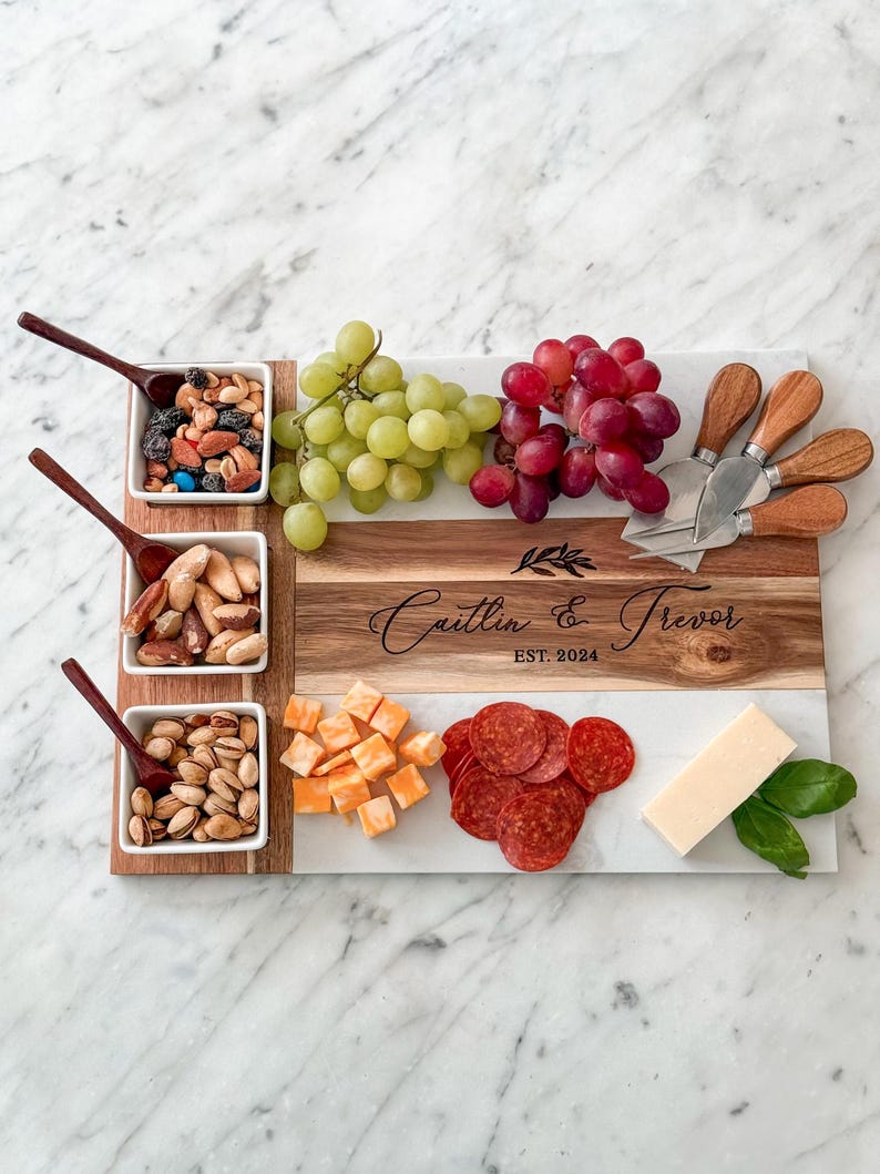 May include: A white marble and wood serving board with four small white bowls filled with snacks. The board has a personalised inscription that reads "Caitlin & Trevor EST. 2024". The board is surrounded by grapes, cheese cubes, pepperoni slices, and basil leaves.