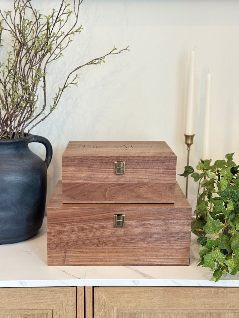May include: Two wooden boxes with brass hinges and latches. The top box has the text "The Allens" engraved on the lid. The boxes are sitting on a white marble surface.