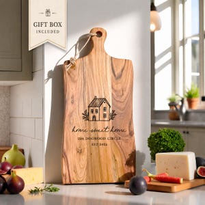 May include: A rectangular wooden cutting board with a handle and a jute string loop. The board features a laser-engraved illustration of a house, the text "home sweet home," "224 DOGWOOD CIRCLE," and "EST. 2025." A gift box tag is attached.