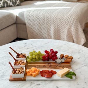 May include: A personalised wooden and marble serving board with a white marble surface and a wooden section with the text "Caitlin & Trevor Est. 2024". The board is filled with various snacks, including grapes, cheese, pepperoni, and nuts.
