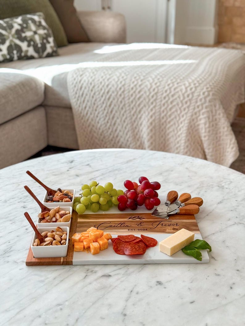 May include: A personalised wooden and marble serving board with a white marble surface and a wooden section with the text "Caitlin & Trevor Est. 2024". The board is filled with various snacks, including grapes, cheese, pepperoni, and nuts.