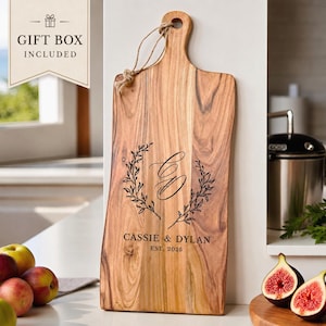 May include: A rectangular wooden serving board with a handle and a jute rope loop. The board features a laser-engraved design of a laurel wreath, the initials CD, the names CASSIE & DYLAN, and the text EST. 2025. A gift box is included.