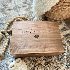 May include: A wooden keepsake box with a heart cutout and the inscription "Tessa Lauren Ninlee June 28, 2024". The box is sitting on a patterned rug with a pair of white baby booties in the foreground.