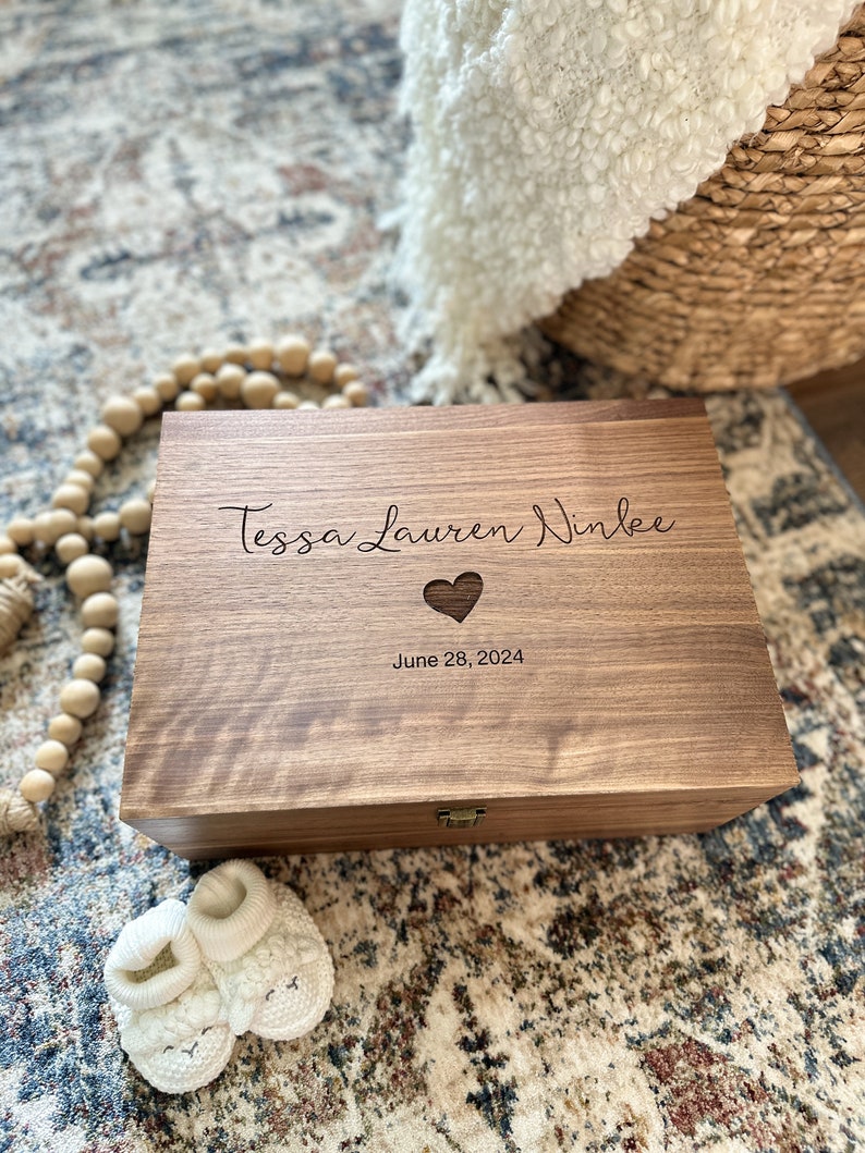 May include: A wooden keepsake box with a heart cutout and the inscription "Tessa Lauren Ninlee June 28, 2024". The box is sitting on a patterned rug with a pair of white baby booties in the foreground.